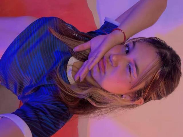 Elodie Gardner knippert met één oog, met haar hand voor haar mond, en draagt een blauw gestreept shirt, een rode armband en een witte kraag, in een paars-blauw licht..