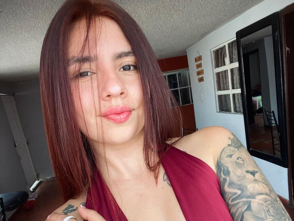 CassieWest, avec des cheveux roux, une peau claire et des lèvres roses, montrant un bras tatoué, vêtue d'un haut marron, avec un léger sourire et une main sur sa poitrine..