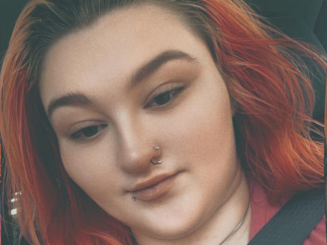 AthenaRosse, con cabello naranja-rojizo, un piercing en la nariz y otro en el labio, lleva puesto el cinturón de seguridad y mira hacia abajo con los labios ligeramente entreabiertos. Tiene un rostro adorable, con pecas y cejas finas..