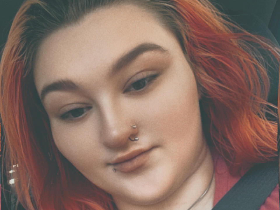 AthenaRosse, con cabello naranja-rojizo, un piercing en la nariz y otro en el labio, lleva puesto el cinturón de seguridad y mira hacia abajo con los labios ligeramente entreabiertos. Tiene un rostro adorable, con pecas y cejas finas..