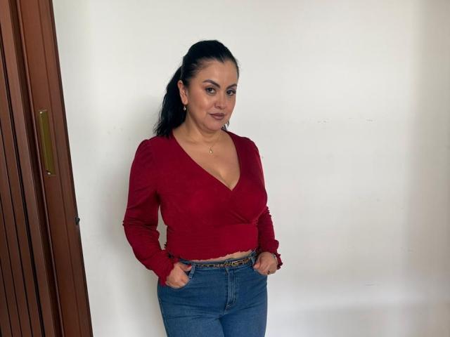 CharlotteDevereaux with dark hair in a ponytail, wearing a red V-neck top, exposing her tits, and blue jeans, standing with hands in pockets, smiling slightly.