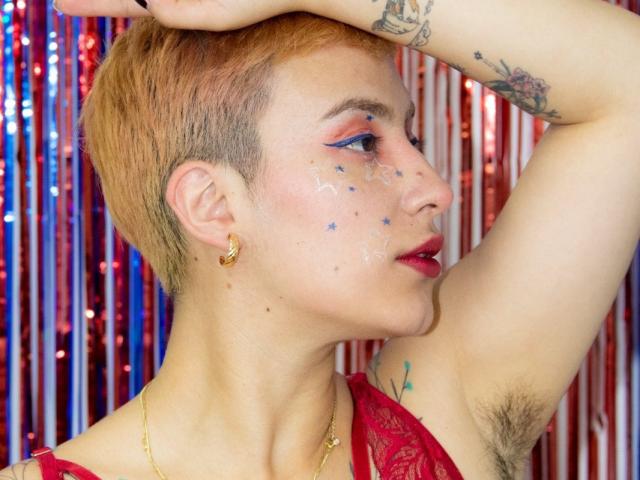 CatsitoS with short blonde hair, blue star makeup on cheek, red lipstick, gold earrings, raised arm, armpit hair, tattoos, red lace top, looking sideways, shiny background.