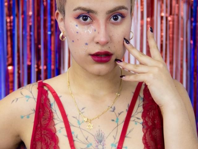 CatsitoS with short hair, blue eye makeup, red lipstick, red lace top, gold necklace, tattoos on chest, painted face, purple nails, gold earrings, touching cheek.