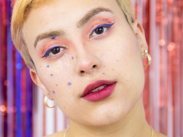 CatsitoS has short blonde hair, blue eyeliner, pink eyeshadow, star stickers on the cheeks, red lipstick, gold hoop earrings, and a slight smile. Skin is fair with some freckles.