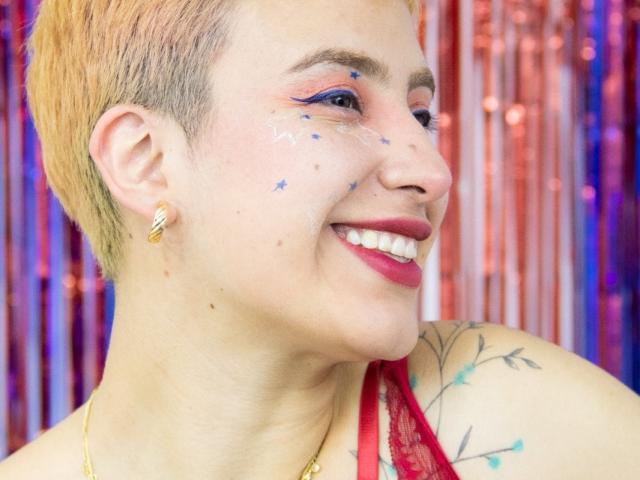 CatsitoS with short, orange-brown hair, blue star makeup on cheeks, red lipstick, and earrings, smiling, showing a tattoo on their right shoulder, wearing a red lace top.