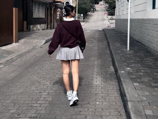 Thalia, vêtue d'un pull violet foncé, d'une jupe blanche et de baskets blanches, marche sur une rue pavée. Elle porte un sac bandoulière noir et a les cheveux attachés en queue de cheval..