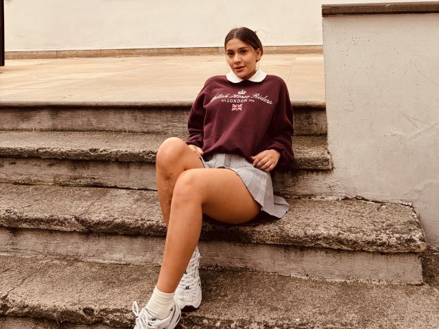 ThaliaAres, zittend op betonnen trappen, draagt een marineblauwe trui, een wit overhemd met kraag, een grijze rok, witte sneakers, met haar blote billen zichtbaar, benen gekruist, lachend, bruin haar, casual..
