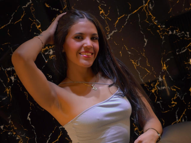VictorieLilieth, con cabello largo y oscuro, sonriendo, vestida con una blusa blanca de un solo hombro con textura vertical, con el brazo levantado, la mano en el cabello, lápiz labial rosa, collar dorado y pulsera, sobre un fondo de mármol oscuro..