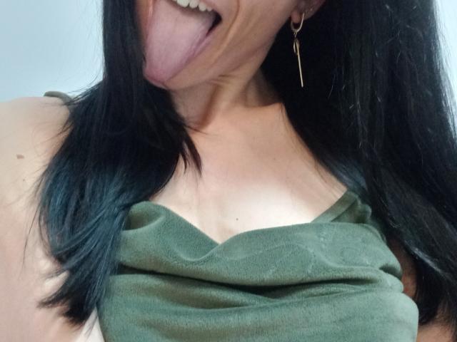 BellaJulls, com cabelo preto, língua de fora, blusa verde, mostrando seios pequenos, usando brincos de ouro, pele clara, com leve decote visível, sorrindo..