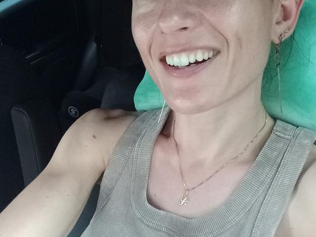 BellaJulls sonriendo con dientes blancos, vistiendo una camiseta gris de tirantes con textura, un collar pequeño con un dije, y un reposacabezas verde. Se ve un asiento de coche. Su brazo izquierdo está levantado y tiene la piel clara..