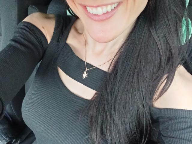 BellaJulls, con cabello largo y negro, sonriendo, vestida con una blusa negra de canalé con un corte en un hombro, y un pequeño collar dorado con una cruz. Sus senos son visibles a través del corte de la blusa..