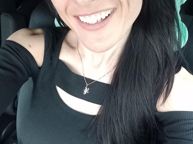 BellaJulls, con cabello negro, sonriendo, vestida con una camiseta negra de canalé con detalles recortados que dejan ver su clavícula, y un collar con un pequeño colgante, sentada en un coche..