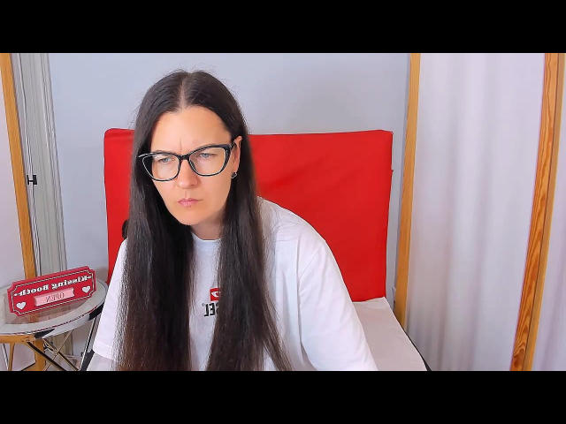 NadiaRose ha lunghi capelli neri, occhiali neri e una camicia bianca con un logo, ed è seduta su una sedia rossa. Guarda leggermente di lato con un'espressione neutra..