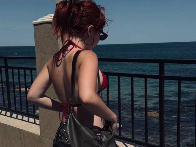 AlexXas with red hair, wearing a red bikini top, showing her back and ass, black leather bag, standing by a metal railing, ocean in the background, sunny day.