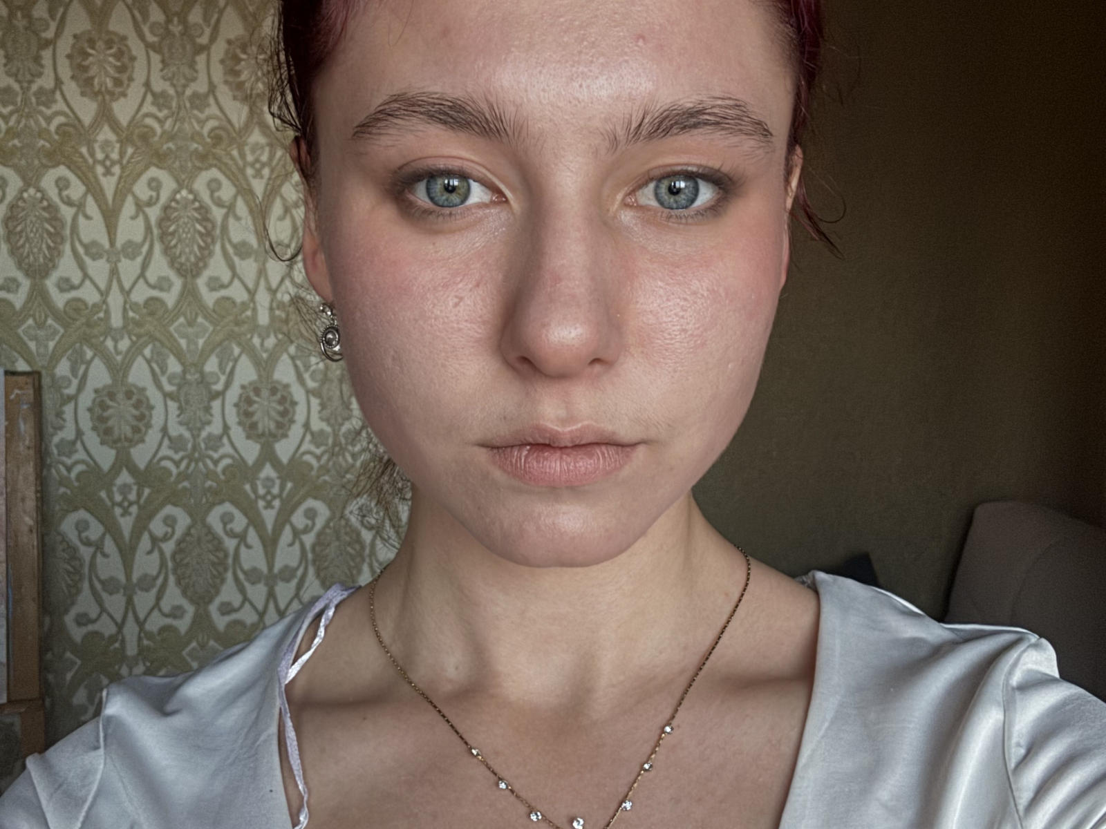 Cristie Razey tem pele clara, olhos verdes e cabelo ruivo, vestindo um vestido branco com um colar delicado. Seu rosto está ligeiramente inchado, com algumas sardas e uma expressão séria..