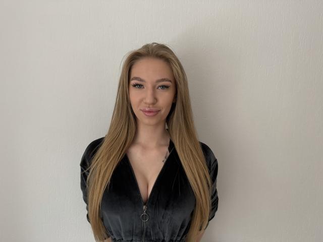 DanielaRecor with long blonde hair, black velvet zip-up, showing big boobs, slight smile, blue eyes, light skin, standing against white wall, looking at camera.