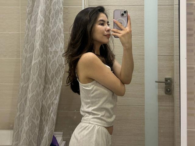 Emerald Crivaro, avec de longs cheveux noirs, de petites seins, portant un débardeur blanc et un pantalon assorti, se prend en photo dans un miroir de salle de bain, souriante, tenant un téléphone rose..