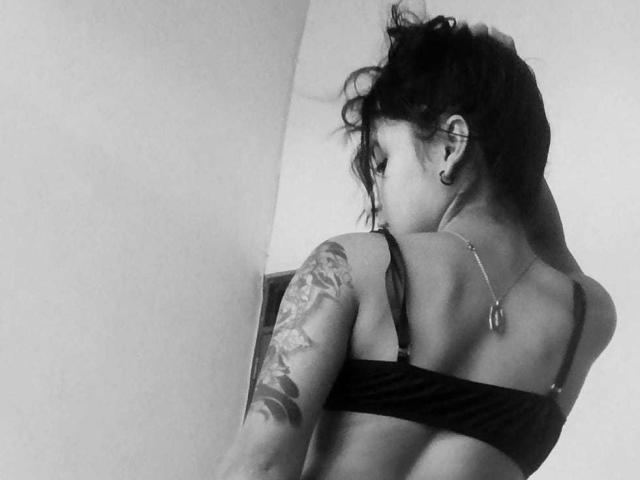 Black-and-white photo of KatheLeclecr with short, messy hair, tattoos on right arm, black bra, back to camera, looking to the side, small dog tattoo on upper back.