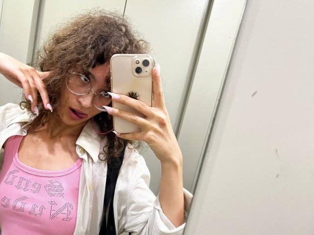 AlyceErway with curly brown hair, wearing glasses and a pink top with glitter, takes a mirror selfie in an elevator. She has pink nails and a white shirt.