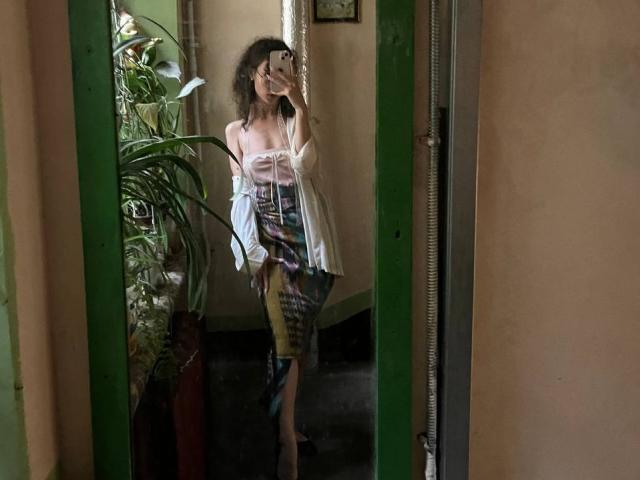 AlyceErway with curly hair, wearing a strapless top, open shirt, and colorful patterned skirt, taking a mirror selfie, showing her tits and ass. Mirror has green frame.