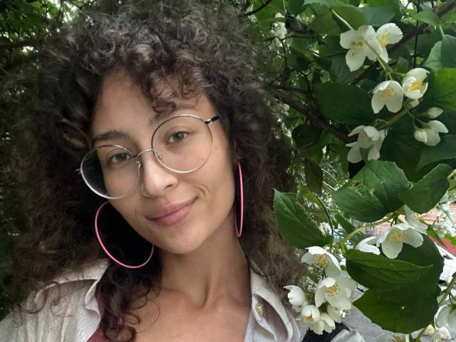 AlyceErway with curly brown hair, green eyes, and pink hoop earrings, wearing glasses and a white shirt, smiling in front of white flowers and green leaves.
