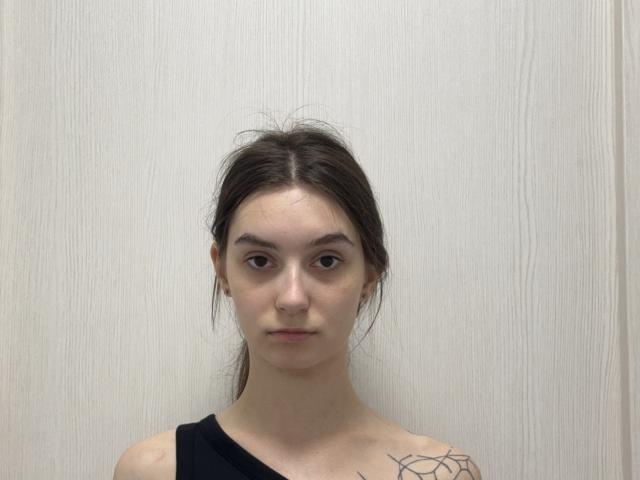 IlaJohal a un visage sérieux, des yeux marron et des cheveux foncés en désordre. Elle porte un haut noir avec un petit tatouage sur son épaule droite, dans une pièce simple..
