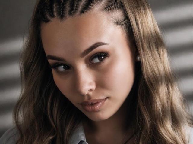 Niferttiti's face with braided crown, long wavy brown hair, dark eyebrows, subtle eyeliner, full lips, and a slight smile. She's looking sideways, wearing a gray shirt.