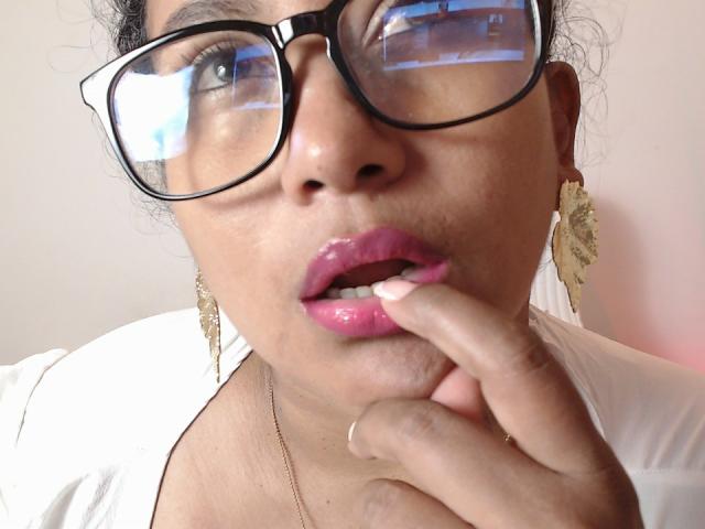YiyaHot69 met glanzende, roze lippen, zwarte brillen en oorbellen van goudfolie, die aan haar vinger zuigt, gekleed in een witte top en een gouden ketting, in een reflecterende selfie..