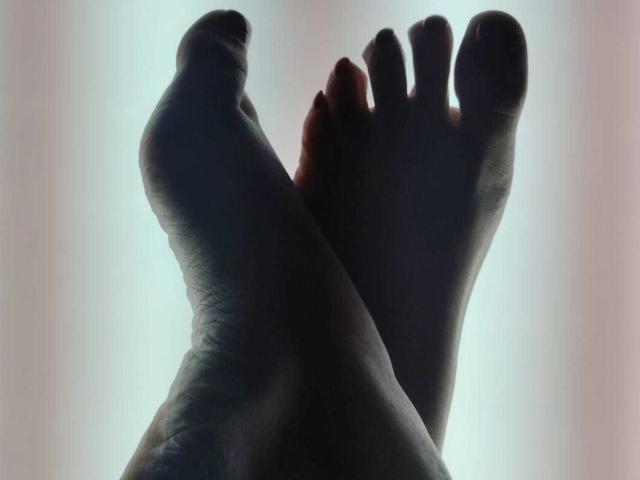 VittoriaI's feet, crossed at the ankles, with dark shadows highlighting their soles and toes against a bright, blurry background. Silhouetted and slightly textured.