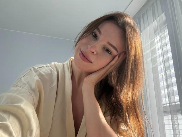 AnnaBlossom, bellissima con i suoi lunghi capelli castani, il vestito bianco e il sorriso dolce, appoggia la testa sulla mano, guardando in basso con un'espressione rilassata e leggermente sensuale..