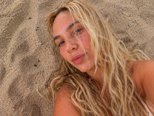 CharlotieRosse, con el cabello rubio despeinado, labios rosados y piel bronceada, acostada en el suelo arenoso, mirando hacia arriba con una expresión relajada, mostrando los hombros desnudos, sin ropa visible ni genitales..