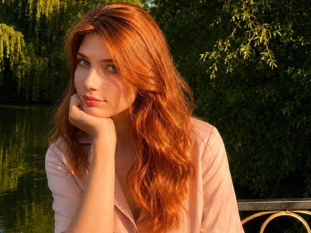 ShellieBessmer, com cabelo ruivo e ondulado, pele clara e olhos verdes, apoiando o queixo na mão, vestindo uma camisa rosa, sentada perto de um curso d'água que reflete a luz..