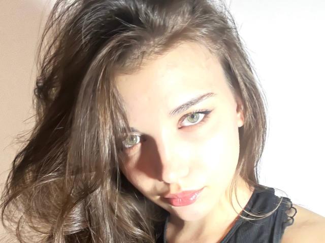 ShellieBessmer, con cabello castaño largo, ojos verdes y lápiz labial rosa, mirando a la cámara con una ligera sonrisa. Lleva una blusa negra. Sus senos son pequeños..