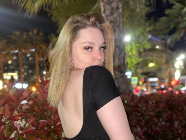 VondaSchlereth mit blonden Haaren, schwarzes, trägerloses Oberteil, das ihren Hintern zeigt, blickt über ihre Schulter, Stadtlichter im Hintergrund, Nacht, Palmenbaum, rote Blätter, leichtes Lächeln, Eyeliner..