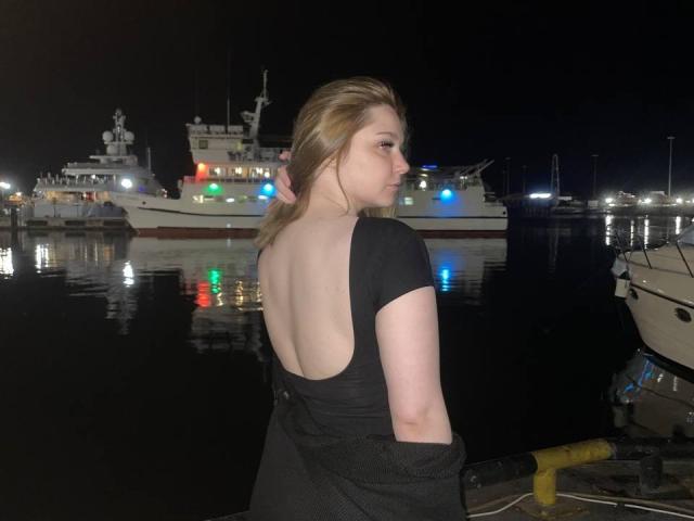 Vonda Schlereth, con cabello rubio, de espaldas a la cámara, vestida con un vestido negro de escote bajo, está de pie junto a un muelle por la noche, con barcos con luces de colores en el fondo..