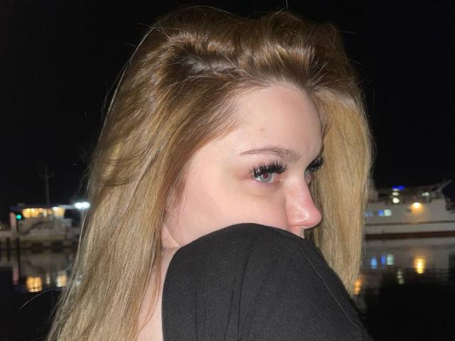 Vonda Schlereth, con lunghi capelli biondi, una maglia nera, occhi azzurri e ciglia lunghe, che si guarda oltre la spalla, ambientazione notturna, riflesso nell'acqua, leggero sorriso, illuminazione soffusa..