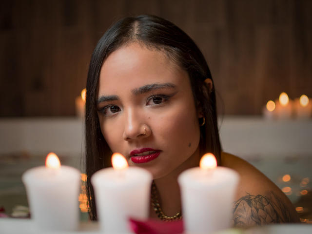 LeaFoster tem cabelo escuro, batom vermelho e um piercing no nariz, e está olhando para velas em uma banheira. Sua tatuagem é visível no ombro direito. Seios parcialmente submersos..