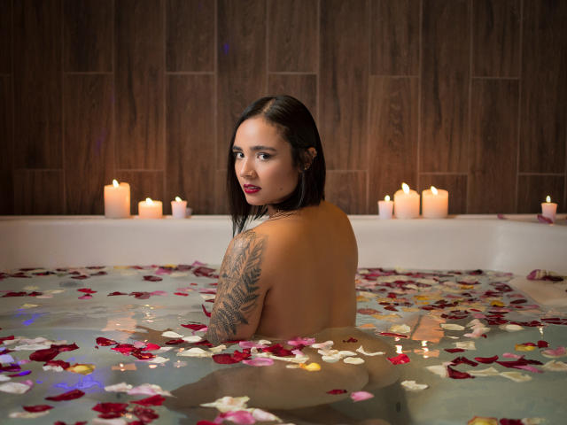 Lea Foster, com seios médios, nua, em uma banheira cheia de água e pétalas de rosa, olhando para trás com um leve sorriso, com velas acesas ao fundo. Ela tem uma tatuagem..