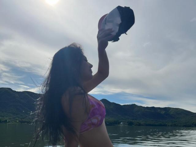 IsabellaaDreams, avec de longs cheveux ondulés, tenant un haut de maillot de bain rose et violet contre un ciel nuageux, debout près d'un lac reflétant, avec des montagnes en arrière-plan..