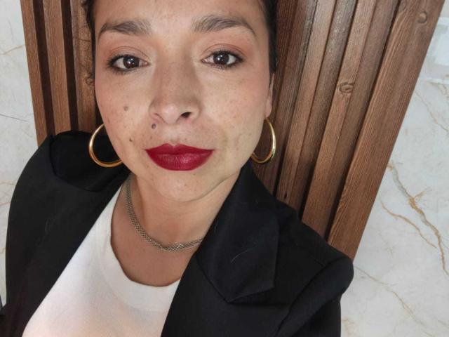 StangelarRivera mit mittlerem Hautton, rotem Lippenstift, Reifenohrringen, einer schwarzen Jacke, einem weißen Hemd, einer Kette und einem Nasenring, steht vor einer vertikalen Holzverkleidung..
