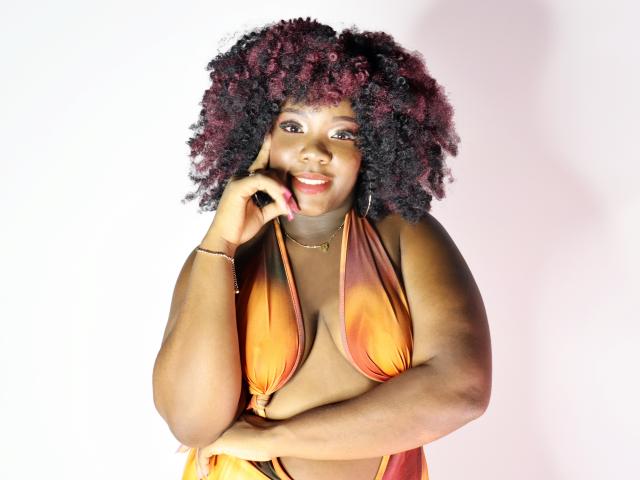 KikiRose tem a pele marrom escura, cabelos cacheados ruivos e pretos, e está vestindo uma blusa cropped laranja e vermelha com estampas tie-dye, que mostra seus seios grandes e arredondados, sorrindo com a mão no rosto..