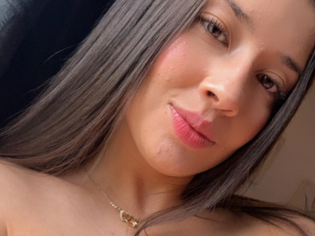 EmmaLombardi has a cute smile with pink lips, brown eyes, and straight dark brown hair, wearing a gold necklace, showing a slight pimple on her right cheek.