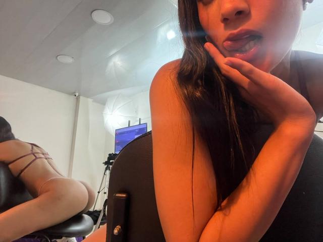 JennTylor, avec de longs cheveux bruns, la langue tirée et un doigt près de la bouche, adoptant une pose séduisante, se tient devant une chaise, le dos tourné vers la caméra, vêtue d'un ensemble de lingerie à fines bretelles, dévoilant ses fesses..
