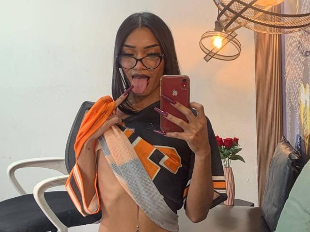 DeboraRaquel met lang zwart haar, een bril en een neusring, met haar tong uit, terwijl ze een handdoek over haar kleine borsten houdt, gekleed in een zwart en oranje sportshirt, een spiegelzelfportret..