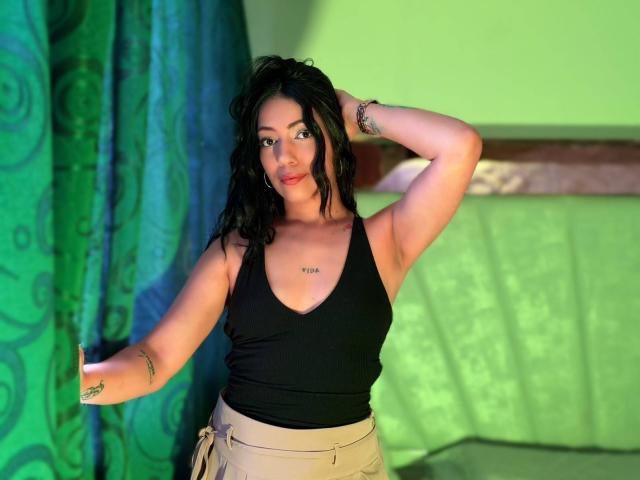 AshleySweetty, avec ses cheveux noirs et ondulés, son débardeur noir et son pantalon beige, debout, une main dans les cheveux, rideau vert, mur vert, petit tatouage sur la poitrine, petit tatouage sur l'avant-bras..