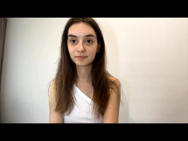 IlaJohal, con lunghi capelli castani, indossa una maglietta bianca con una spalla scoperta, ha un'espressione neutra ed è al centro dell'inquadratura, con una parete bianca semplice dietro di lei..
