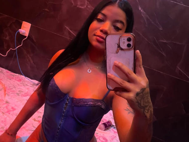 Tattuada69, avec de longs cheveux noirs, portant un corset bleu à lacets, tenant un téléphone rose, montrant des tatouages sur son bras droit, et un collier, dans une pièce faiblement éclairée..