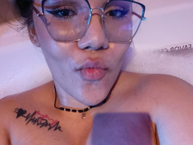 KennyRousse, avec des lunettes, des lèvres boudeuses et un collier noir, arborant un tatouage sur la poitrine représentant des lignes de battements cardiaques et un petit cœur, tenant un téléphone dans un bain moussant..