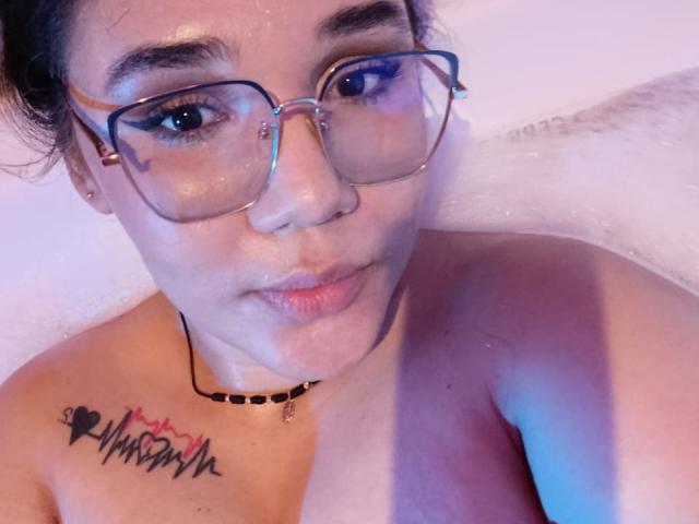 KennyRousse, naakt bovenlichaam, met een bril, lichte huid, subtiele make-up, kleine neusring, zwarte choker, tatoeage boven het linkerborst, roze-blauwe badwater, lichtjes open lippen, nat haar, subtiele glimlach..