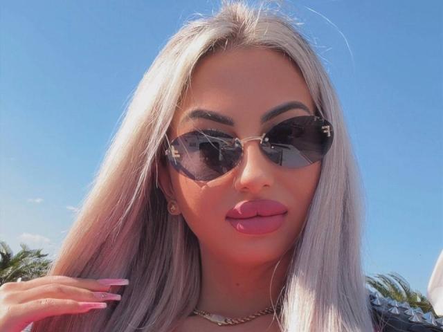 CelineDyor met lang, platina blond haar, die ronde, donkere zonnebrillen draagt, roze lippenstift en een gouden ketting, waarbij haar roze, verzorgde nagels te zien zijn tegen een helderblauwe lucht..